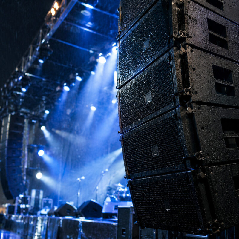 Polyurea-coated speaker cabinets at an outdoor concert in rain, showing waterproof protection for pro audio touring equipment